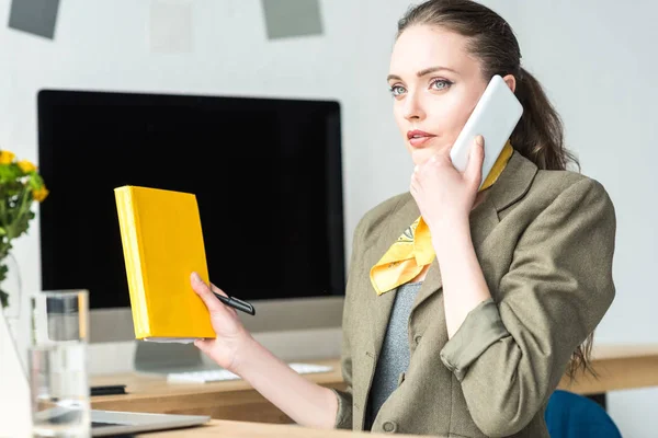 Bela mulher de negócios elegante segurando livro e falando por smartphone no local de trabalho — Fotografia de Stock