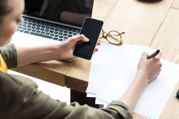 Plan recadré de femme d'affaires en utilisant un smartphone avec écran vierge et en prenant des notes sur le lieu de travail — Photo de stock