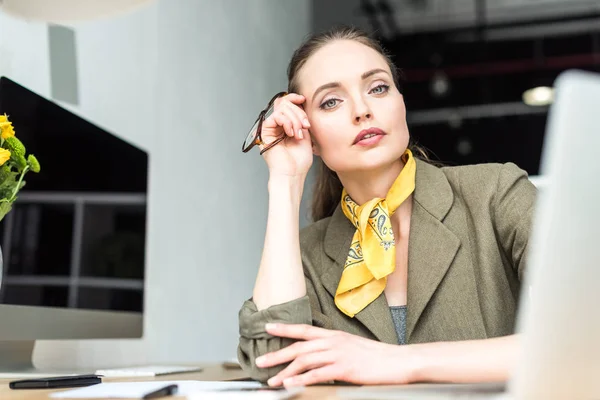 Portrait of beautiful businesswoman holding eyeglasses and looking at camera at workplace — Stock Photo