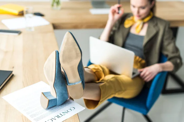 Selektiver Fokus der Geschäftsfrau, die Laptop benutzt und mit Beinen am Tisch mit Vertrag sitzt — Stockfoto