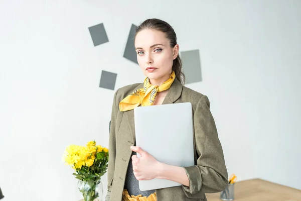 Beautiful stylish businesswoman holding laptop and looking at camera in office — Stock Photo