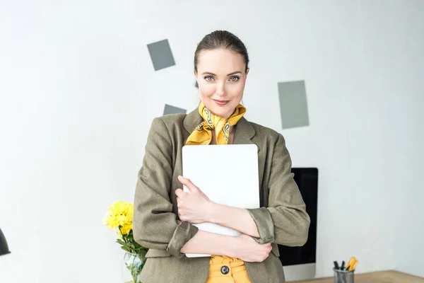 Bella donna d'affari elegante che tiene il computer portatile e sorride alla fotocamera in ufficio — Foto stock