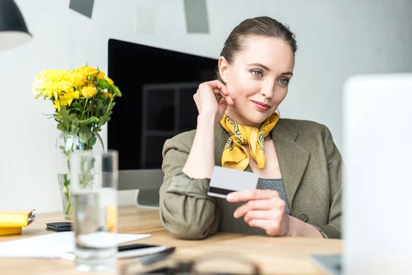 Sorridente mulher de negócios elegante segurando cartão de crédito e olhando para laptop no escritório — Fotografia de Stock