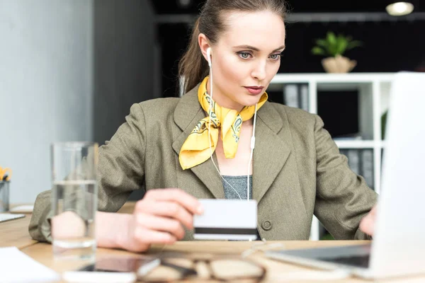 Стильная деловая женщина в наушниках с кредитной картой и ноутбуком в офисе — стоковое фото