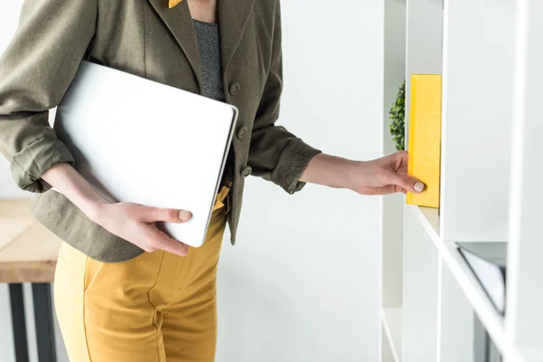 Cropped shot of businesswoman holding laptop and taking book from shelf in office — Stock Photo