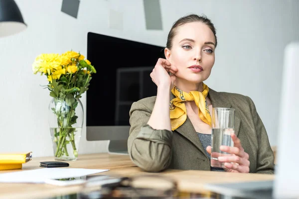 Beautiful pensive businesswoman holding glass of water and looking away in office — Stock Photo