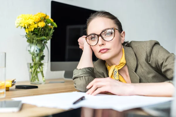 Mujer de negocios pensativa en anteojos apoyados en la mesa y mirando hacia otro lado en la oficina - foto de stock