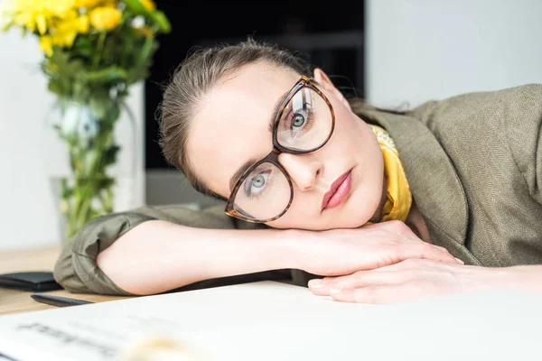Porträt der schönen Geschäftsfrau mit Brille, die in die Kamera blickt, während sie sich im Büro an den Tisch lehnt — Stockfoto