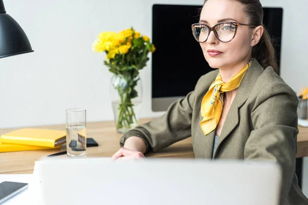 Atractiva mujer de negocios sonriente en anteojos mirando hacia otro lado mientras está sentada en el lugar de trabajo — Stock Photo