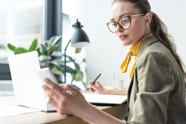 Attraktive Geschäftsfrau in Brille mit Smartphone und Notizen im Büro — Stockfoto