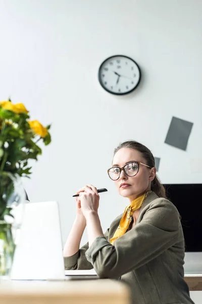 Beautiful middle aged businesswoman in eyeglasses using laptop and looking at camera in office — Stock Photo