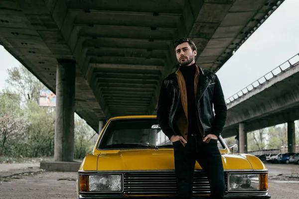 Handsome stylish young man in leather jacket standing with hands in pockets near yellow retro automobile — Stock Photo