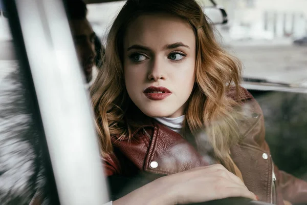 Schöne junge Frau in Lederjacke schaut weg, während sie im Auto sitzt — Stockfoto