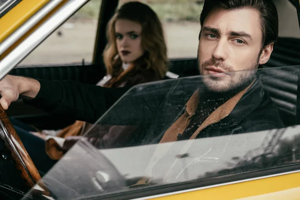 Guapo joven con estilo mirando a la cámara mientras está sentado en el coche con hermosa novia — Stock Photo