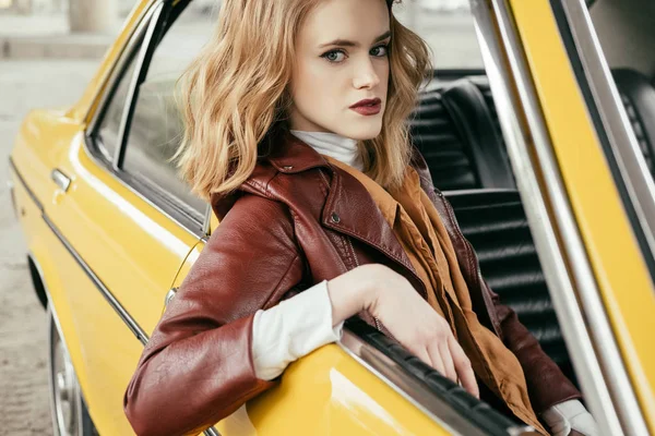 Красивая стильная девушка в кожаной куртке сидит в старомодной машине и смотрит в камеру — стоковое фото