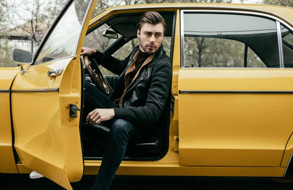 Красивий стильний молодий чоловік у шкіряній куртці сидить у жовтій класичній машині і дивиться вбік — стокове фото