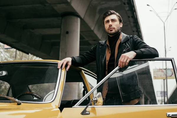 Vista de ángulo bajo de hombre elegante guapo mirando hacia otro lado, mientras que de pie cerca de coche clásico amarillo - foto de stock