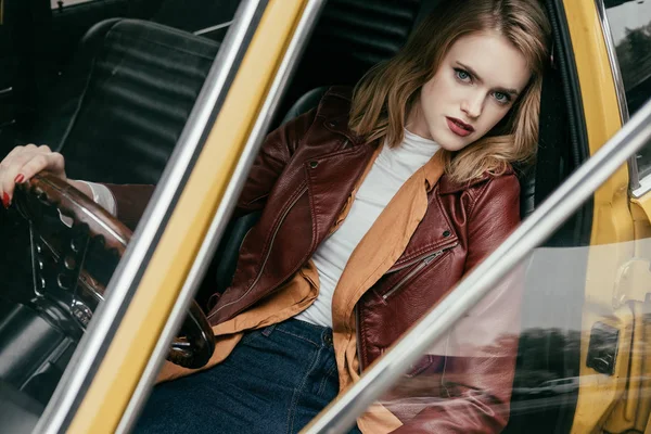 Vista de ángulo alto de hermosa chica en chaqueta de cuero sentado en coche retro y mirando a la cámara - foto de stock