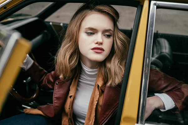 Belle jeune femme élégante assise dans une voiture à l'ancienne et regardant loin — Photo de stock