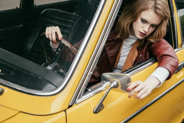 Vista de ángulo alto de hermosa mujer joven con estilo en chaqueta de cuero sentado en coche vintage amarillo - foto de stock