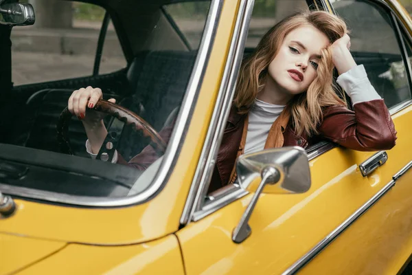 Jovem atraente olhando para a câmera e segurando o volante enquanto sentado no carro retro — Fotografia de Stock