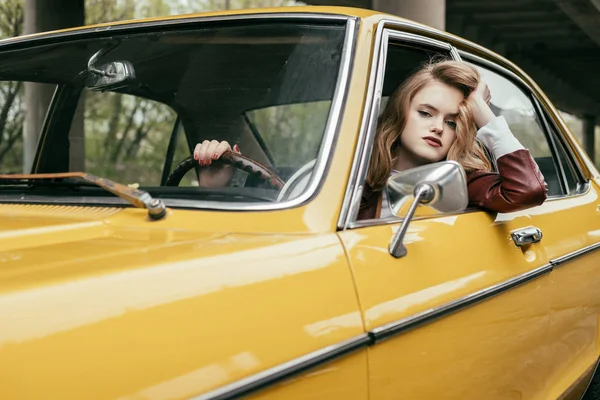 Belle jeune femme regardant la caméra et tenant le volant tout en étant assis dans une voiture vintage — Photo de stock