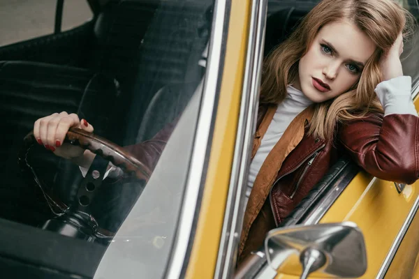 Высокий угол обзора красивой девушки, смотрящей на камеру и держащей руль, сидя в классическом автомобиле — стоковое фото