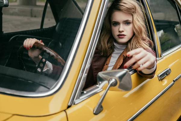 Schöne junge Frau in Lederjacke blickt in die Kamera, während sie im gelben Retro-Auto sitzt — Stockfoto