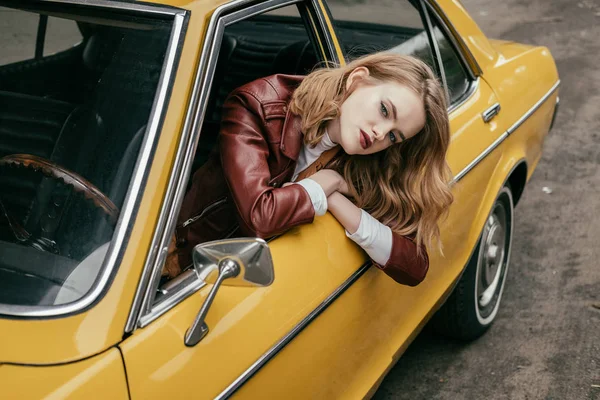 Високий кут зору красива стильна дівчина дивиться на камеру через вікно жовтого класичного автомобіля — стокове фото