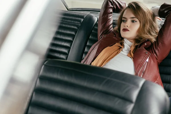 Foco seletivo de menina bonita sensual em jaqueta de couro sentado no carro e olhando para longe — Fotografia de Stock