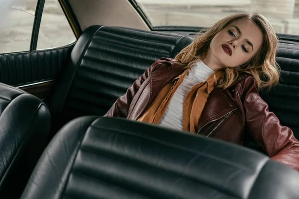 Bela menina sensual em jaqueta de couro sentado no carro e olhando para longe — Fotografia de Stock