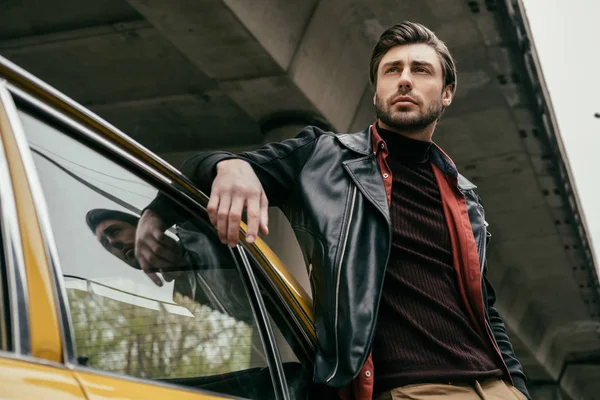 Vista de ángulo bajo del hombre guapo apoyado en el coche y mirando hacia otro lado - foto de stock