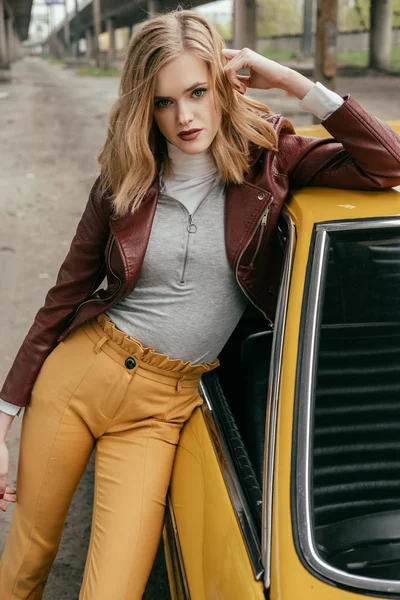 Beautiful stylish young woman leaning at yellow car and looking at camera — Stock Photo