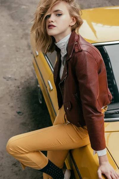 Élégante jeune femme en veste en cuir regardant la caméra tout en étant assis sur une voiture vintage jaune — Photo de stock