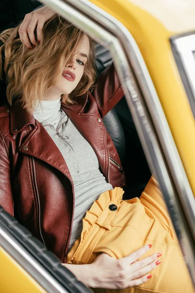 Vista de alto ângulo de menina elegante em jaqueta de couro sentado no carro vintage e olhando para a câmera — Fotografia de Stock