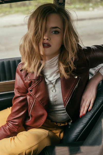 Chica seductora en chaqueta de cuero mirando a la cámara mientras está sentado en el coche - foto de stock