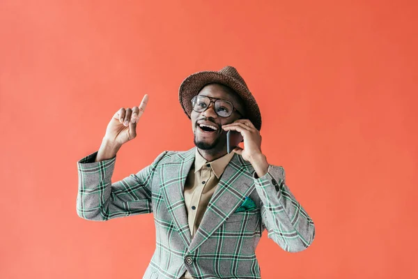 Hombre afroamericano emocionado hablando en el teléfono inteligente y apuntando hacia arriba con idea, aislado en rojo — Stock Photo