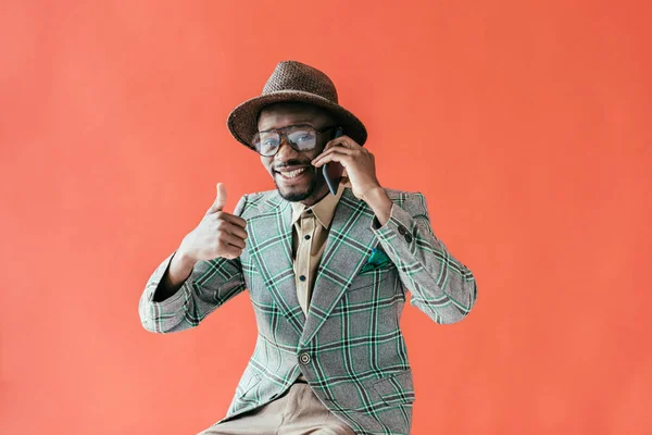 Hombre afroamericano de moda con el pulgar hacia arriba hablando en el teléfono inteligente, aislado en rojo - foto de stock