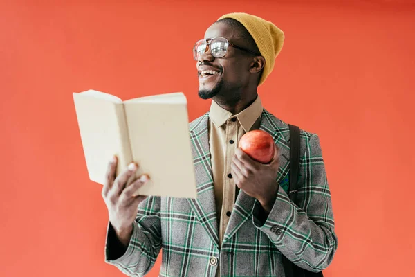 Hombre americano africano risueño elegante con el libro y la manzana, aislado en rojo - foto de stock