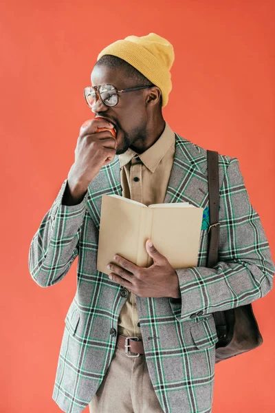 Hombre afroamericano guapo con libro comer manzana, aislado en rojo - foto de stock