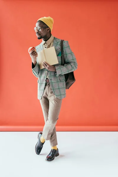 Homme afro-américain à la mode posant avec livre et pomme, sur rouge — Photo de stock