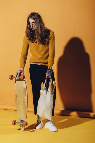 Stylish skateboarder holding longboard and bag with food, on yellow — Stock Photo