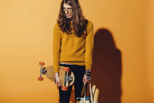 Bel pattinatore che tiene longboard e borsa con cibo, su giallo — Foto stock