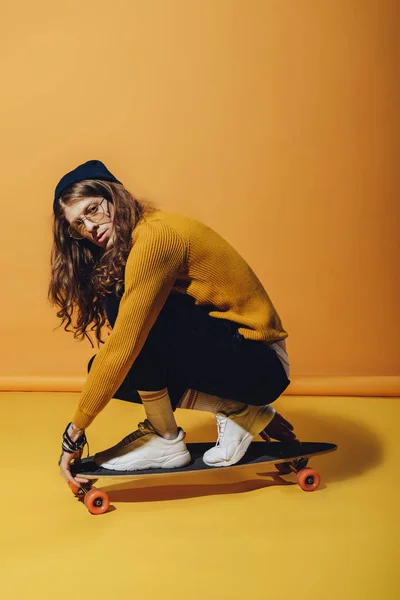Stylish skateboarder with long hair sitting on longboard, on yellow — Stock Photo