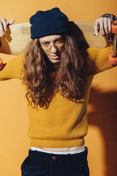 Stylish skater with long hair holding longboard, on yellow — Stock Photo