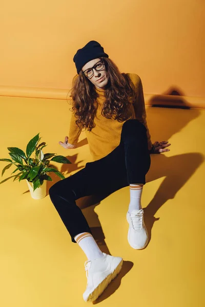 Homem elegante com cabelos longos sentado perto de planta de sala, em amarelo — Fotografia de Stock