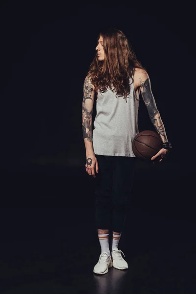 Tattooed man with long hair holding basketball ball, on black — Stock Photo