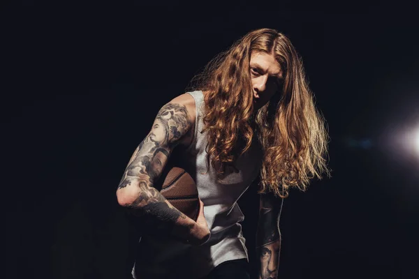 Hombre tatuado con el pelo largo jugando baloncesto, aislado en negro - foto de stock