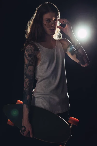 Tattooed skateboarder with long hair posing with longboard, on black with backlit — Stock Photo