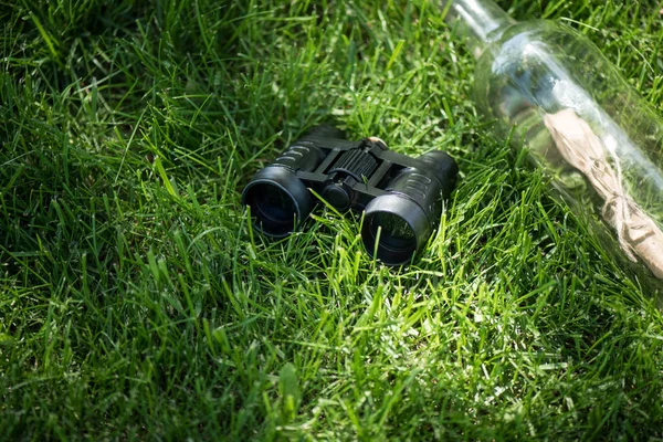 Vista de perto de binóculos e papel em garrafa de vidro dispostos no gramado verde — Fotografia de Stock